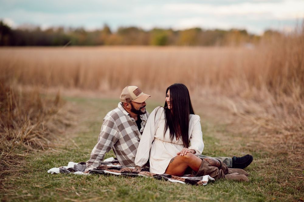 A couple sits together on a plaid blanket in a golden autumn field during sunset.