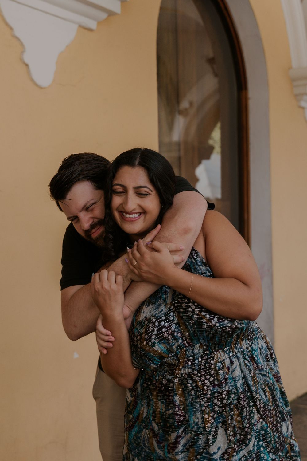 A couple shares a joyful moment together, embracing and laughing in front of an arched doorway with beige walls.
