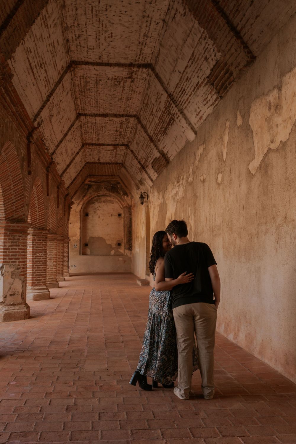 Two people stand close together in a dimly lit brick archway corridor inside a historic building.