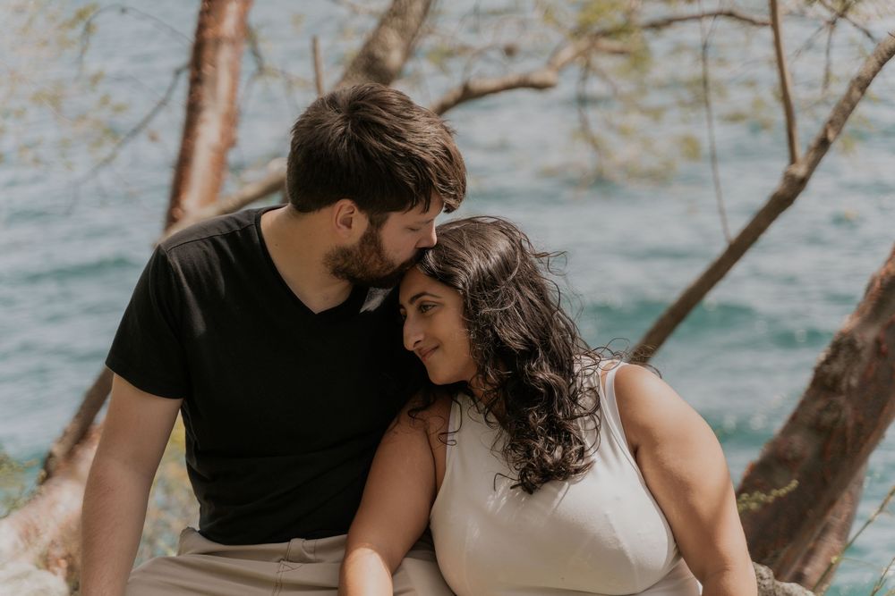 Romantic couple embraces near water's edge with sun-dappled trees and ocean in background.