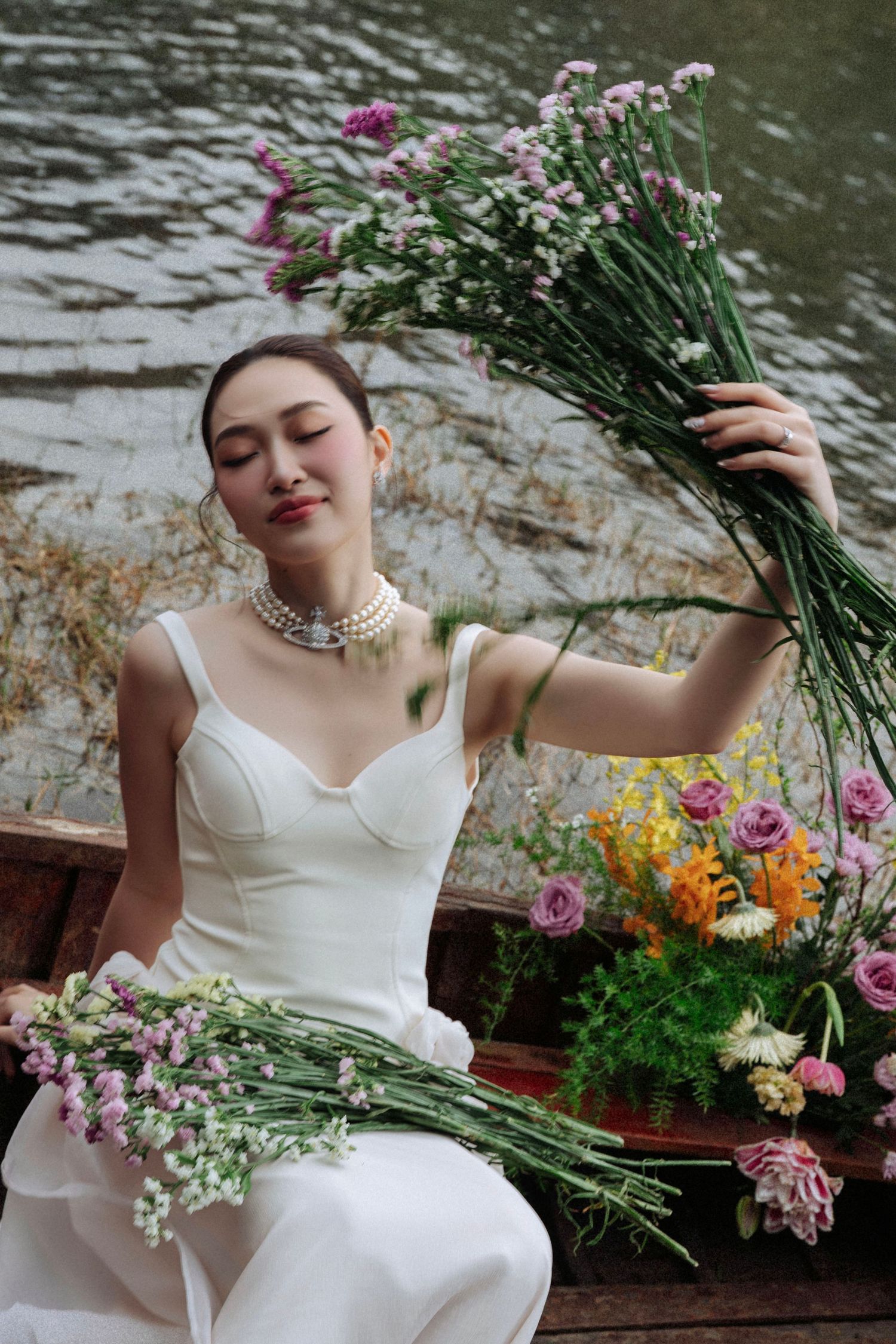 Joyful bride in white gown with pearl necklace raising a wildflower bouquet on a flower-filled boat.