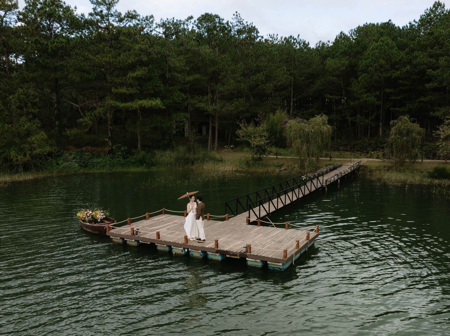 Bride in white dress standing on a wooden dock over a serene green lake surrounded by lush forest.