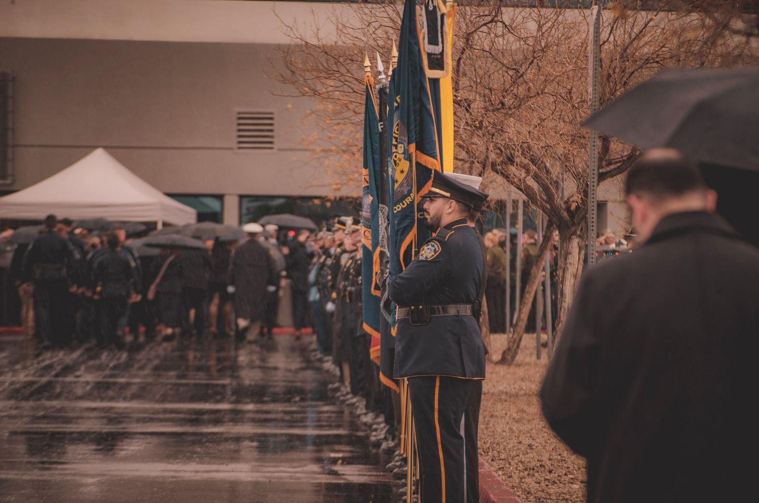 Police honor guard stands at attention holding flags during a rainy outdoor ceremony.