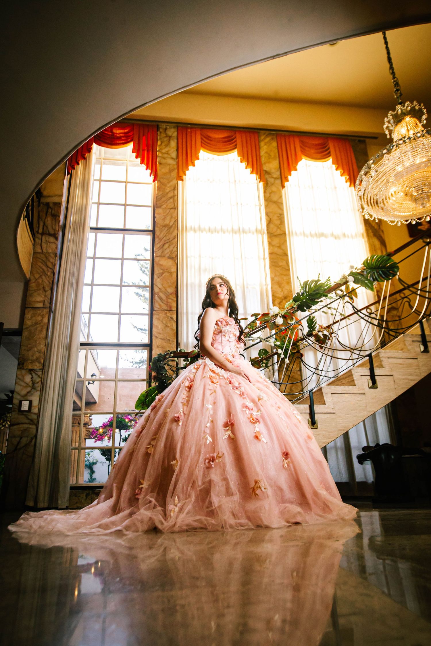 A stunning pink ballgown dress glows in front of tall windows with gold curtains and a grand crystal chandelier.