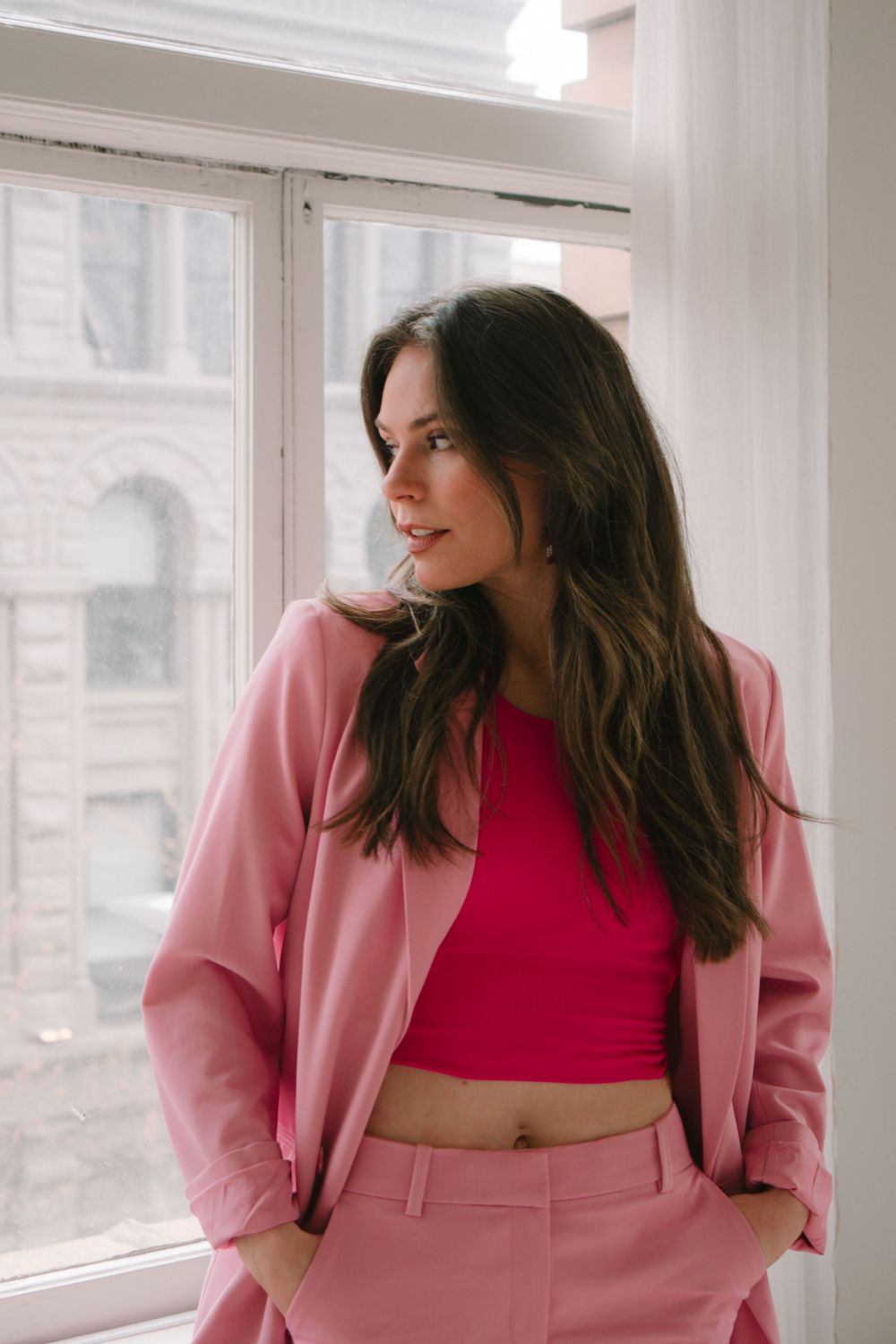 Person in pink suit and red top poses by window in modern urban setting.