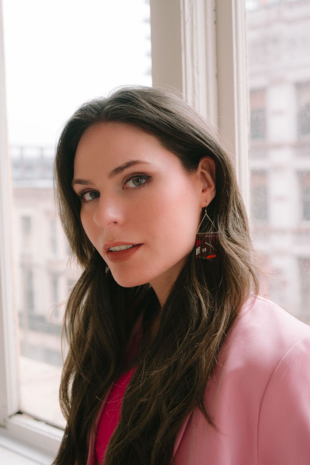 Person in pink blazer and red top poses by bright window.