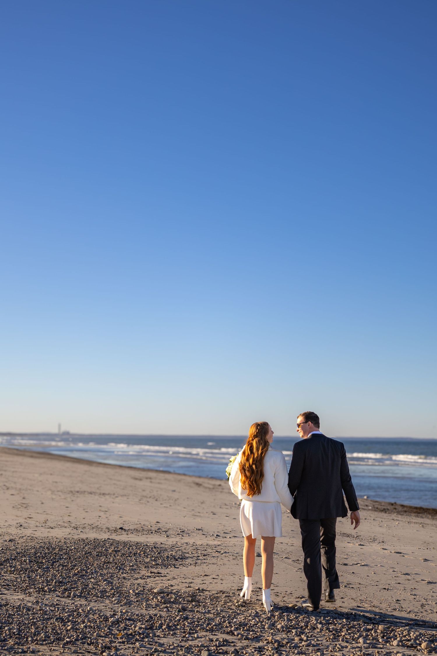 Couple walking along a serene beach shoreline at sunset wearing formal attire.