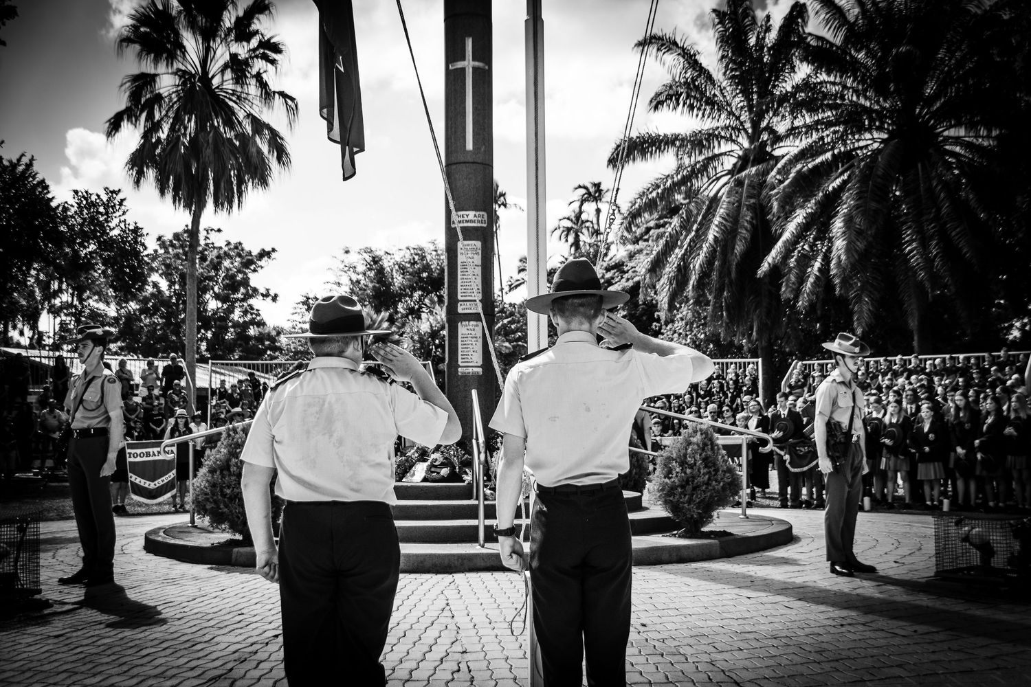 Two figures stand at a street light pole in black and white with palm trees and clothing vendors in the background.