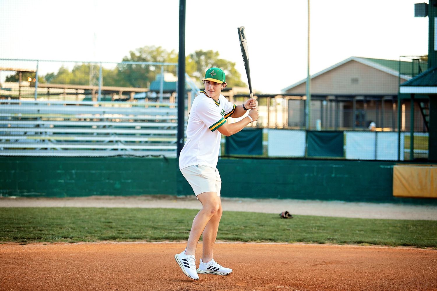 Baseball player practicing swing form on dirt infield with bleachers in background.