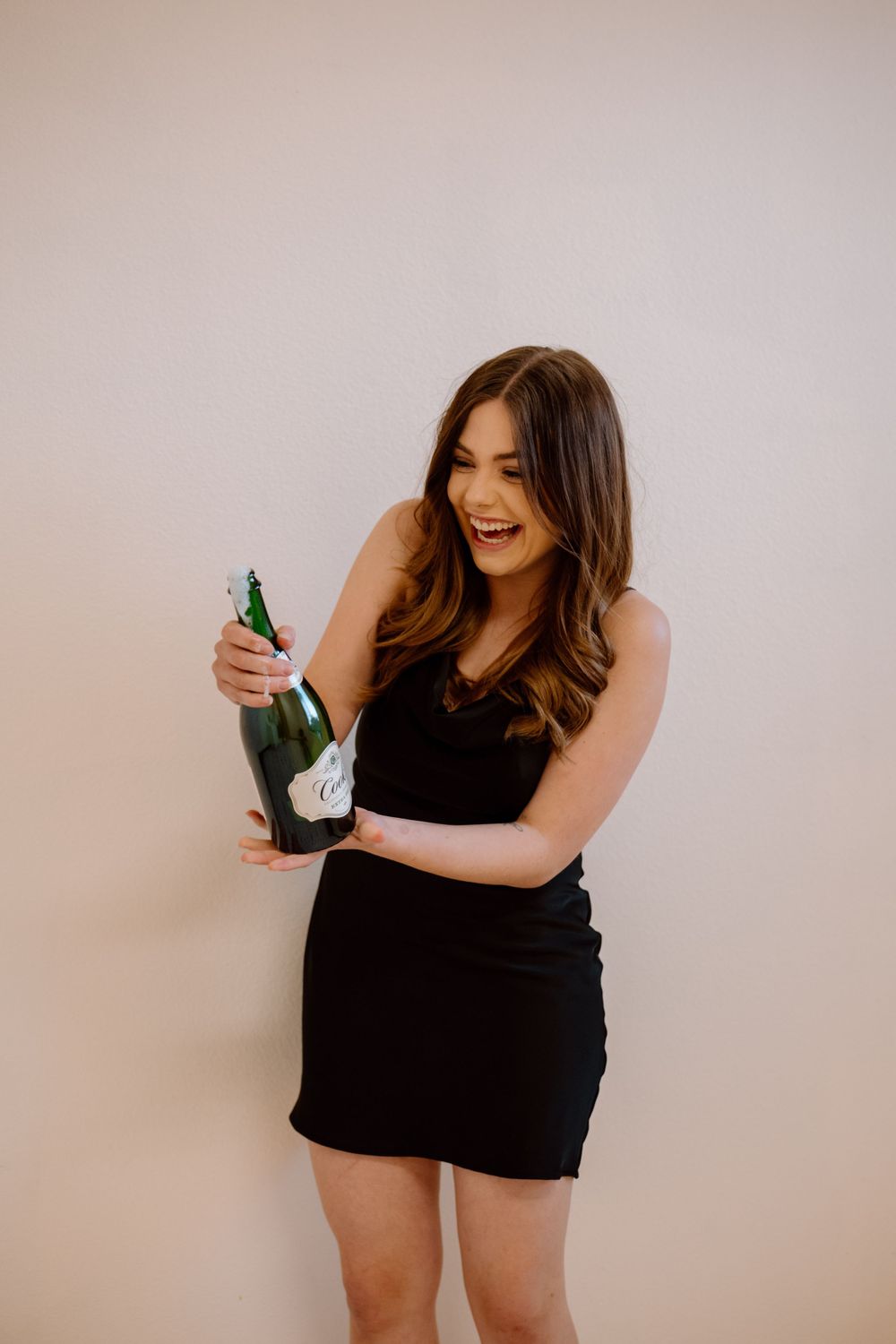 A person in a black dress celebrates with a champagne bottle against a beige wall.