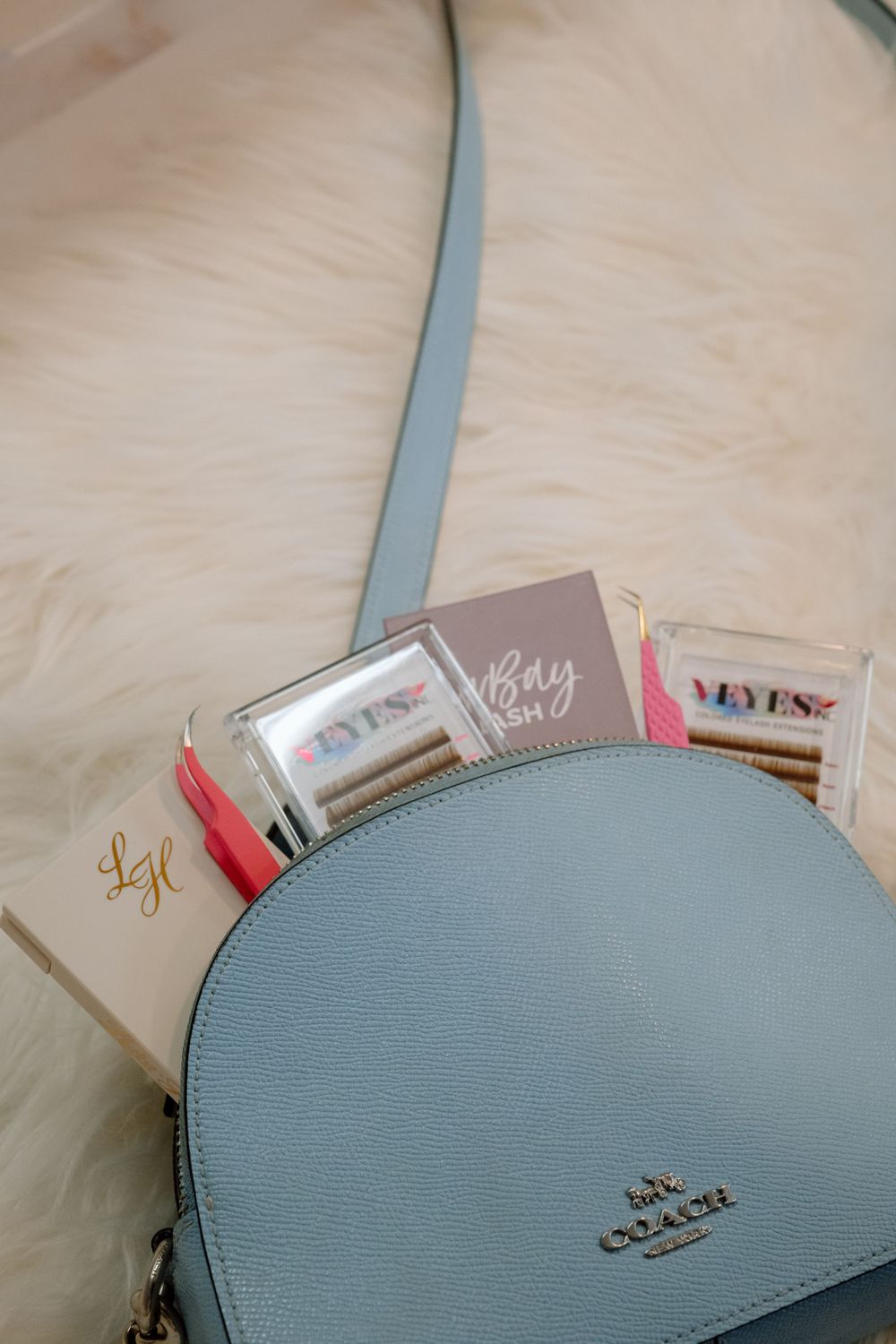 A light blue Coach half-moon purse with makeup products displayed on a cream surface.