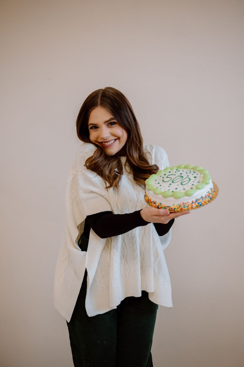 A person holds a decorated cake while wearing a white blouse and dark pants against a neutral background.