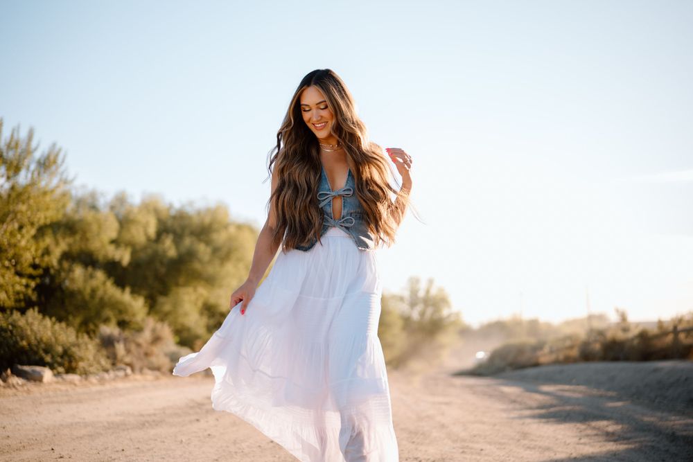 Person in denim top and flowing white skirt twirls on a dirt path at sunset.