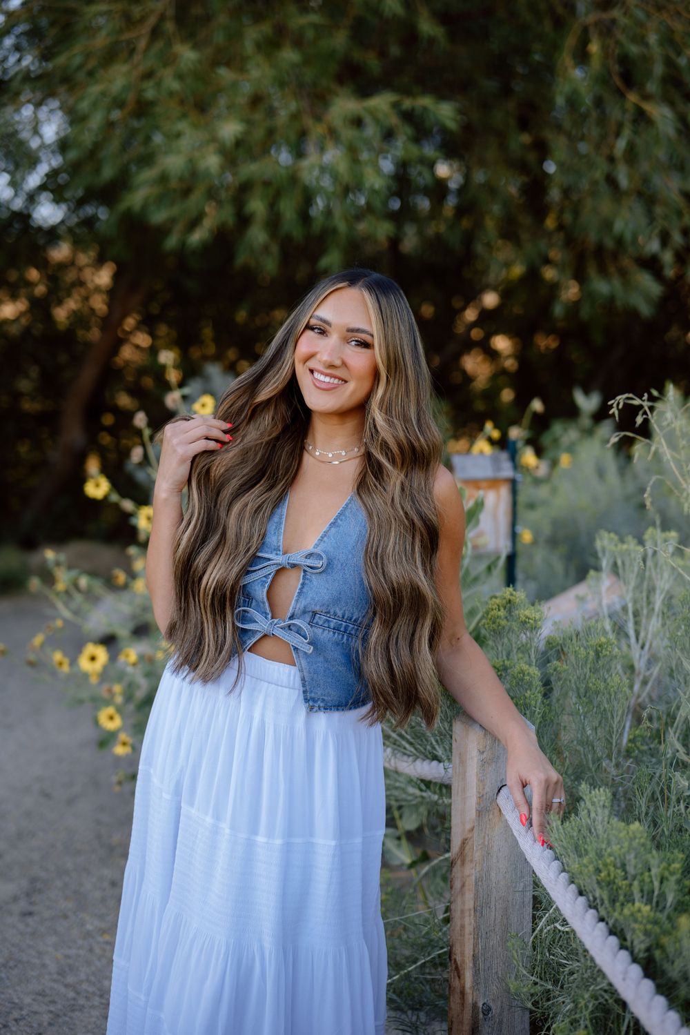 Person in denim crop top and white maxi skirt poses along garden path with yellow wildflowers.