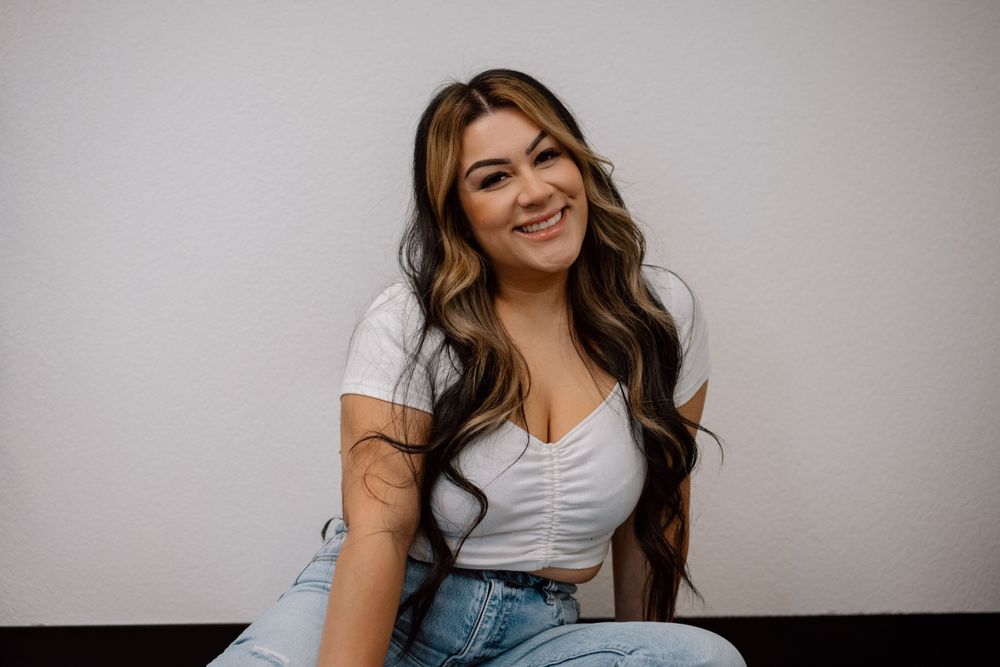 Person in white top and jeans sits casually against neutral background while smiling.