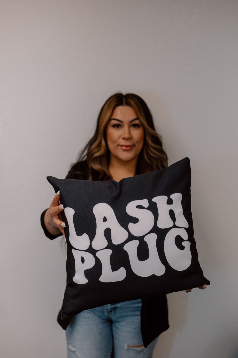 Person holding black decorative pillow with 'LASH PLUG' text in casual attire.