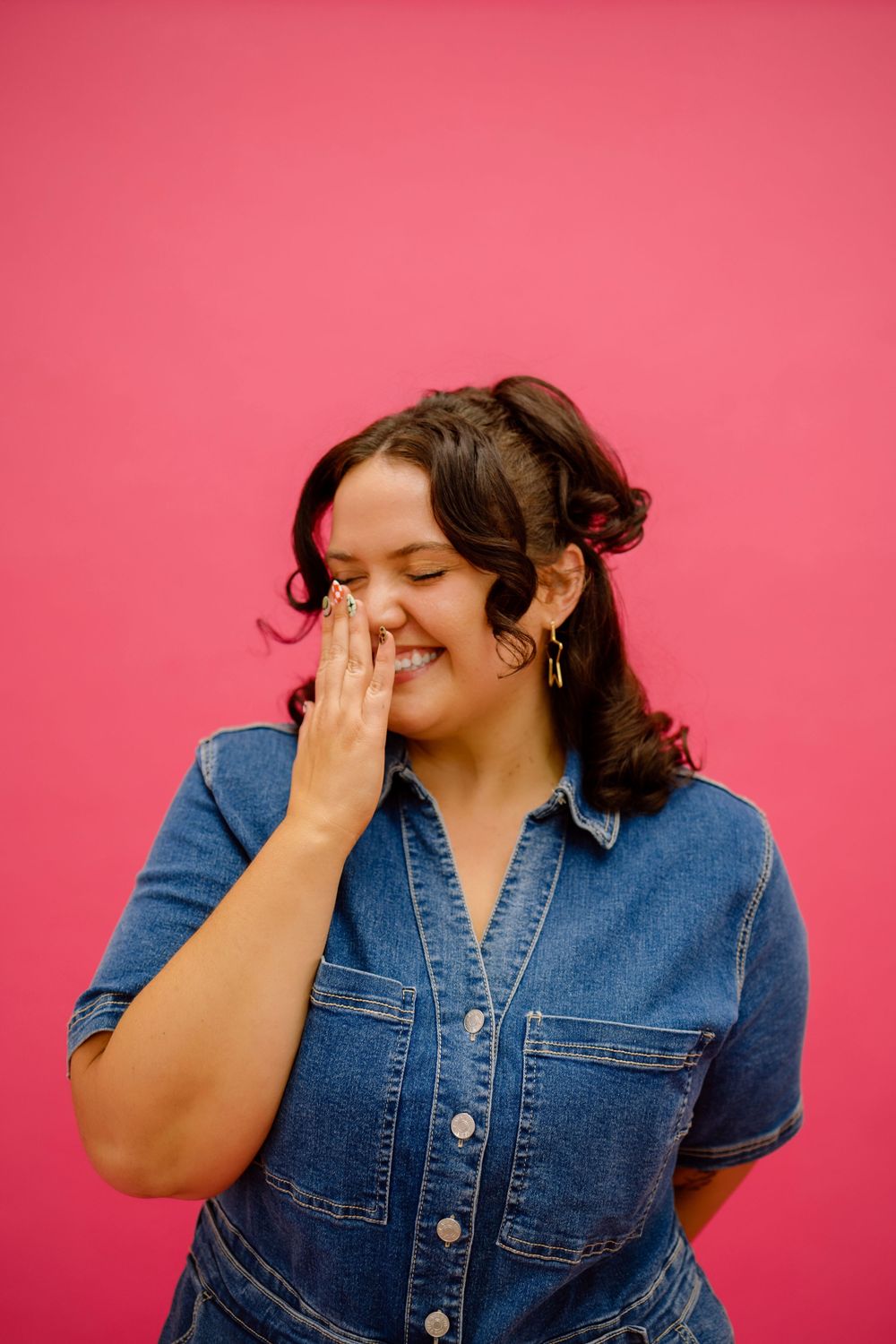 A person in a denim shirt laughs while covering their face against a bright pink background.