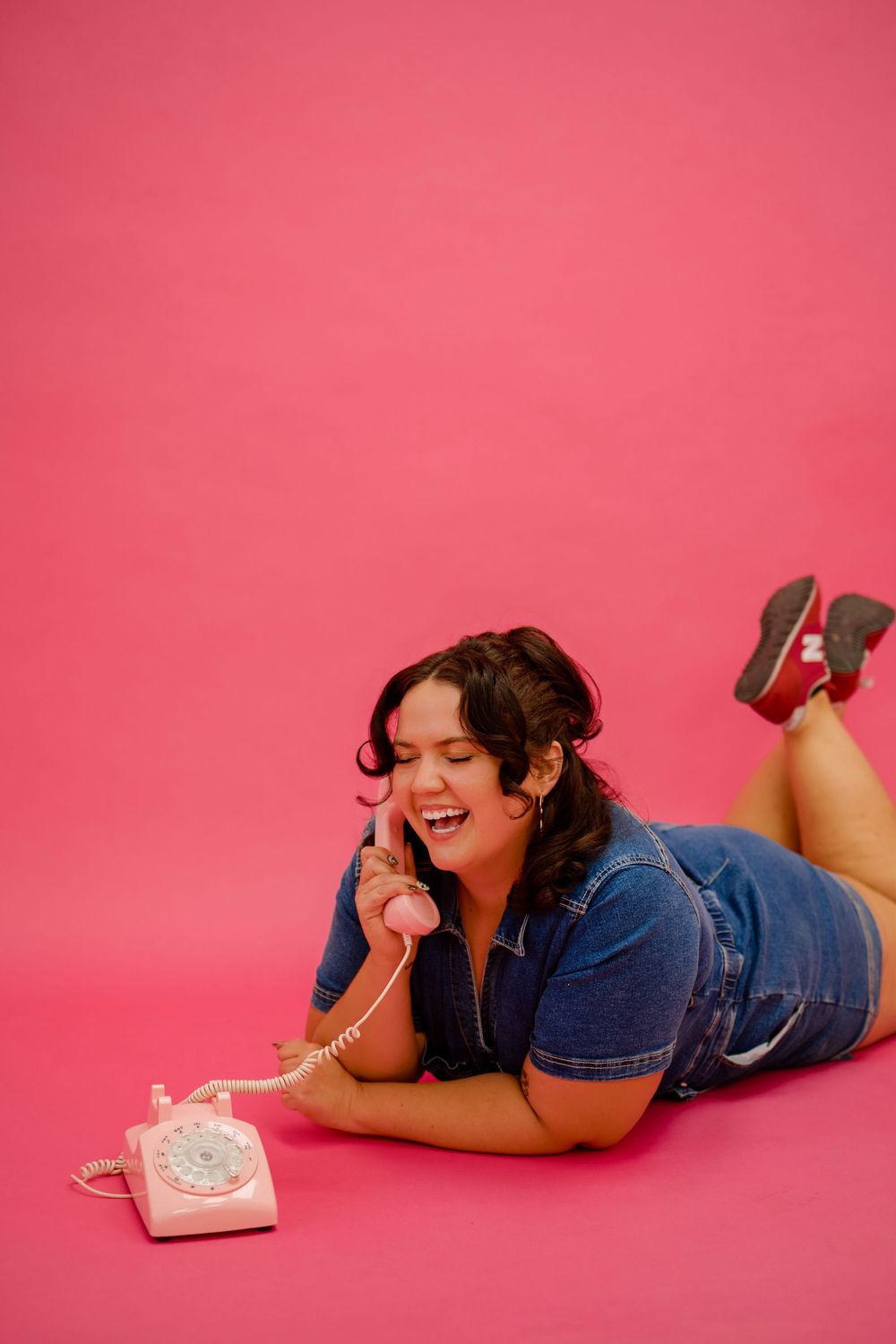 Someone in denim lying on pink background with vintage rotary telephone in playful retro photoshoot.