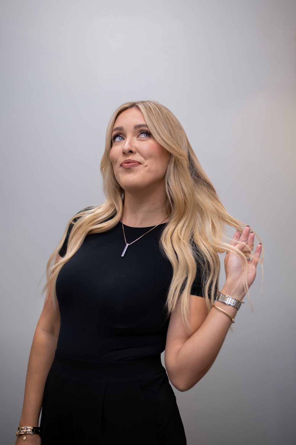 Woman in black dress with long blonde wavy hair poses against grey background with raised hand.