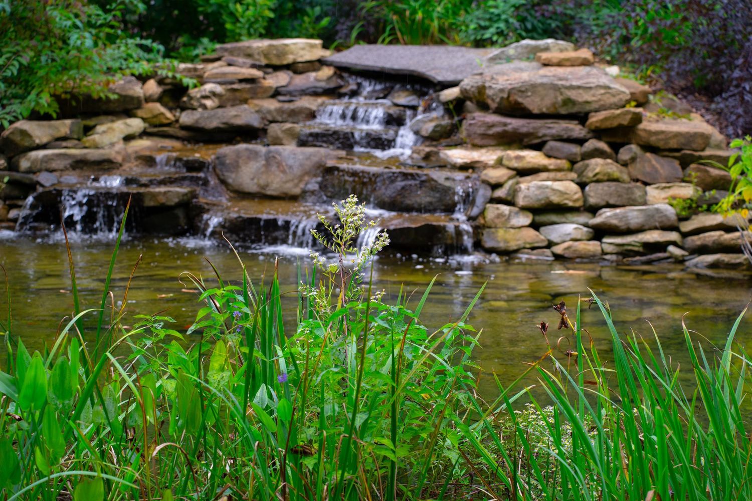 Serene garden waterfall flowing over stacked rocks into a green pond.