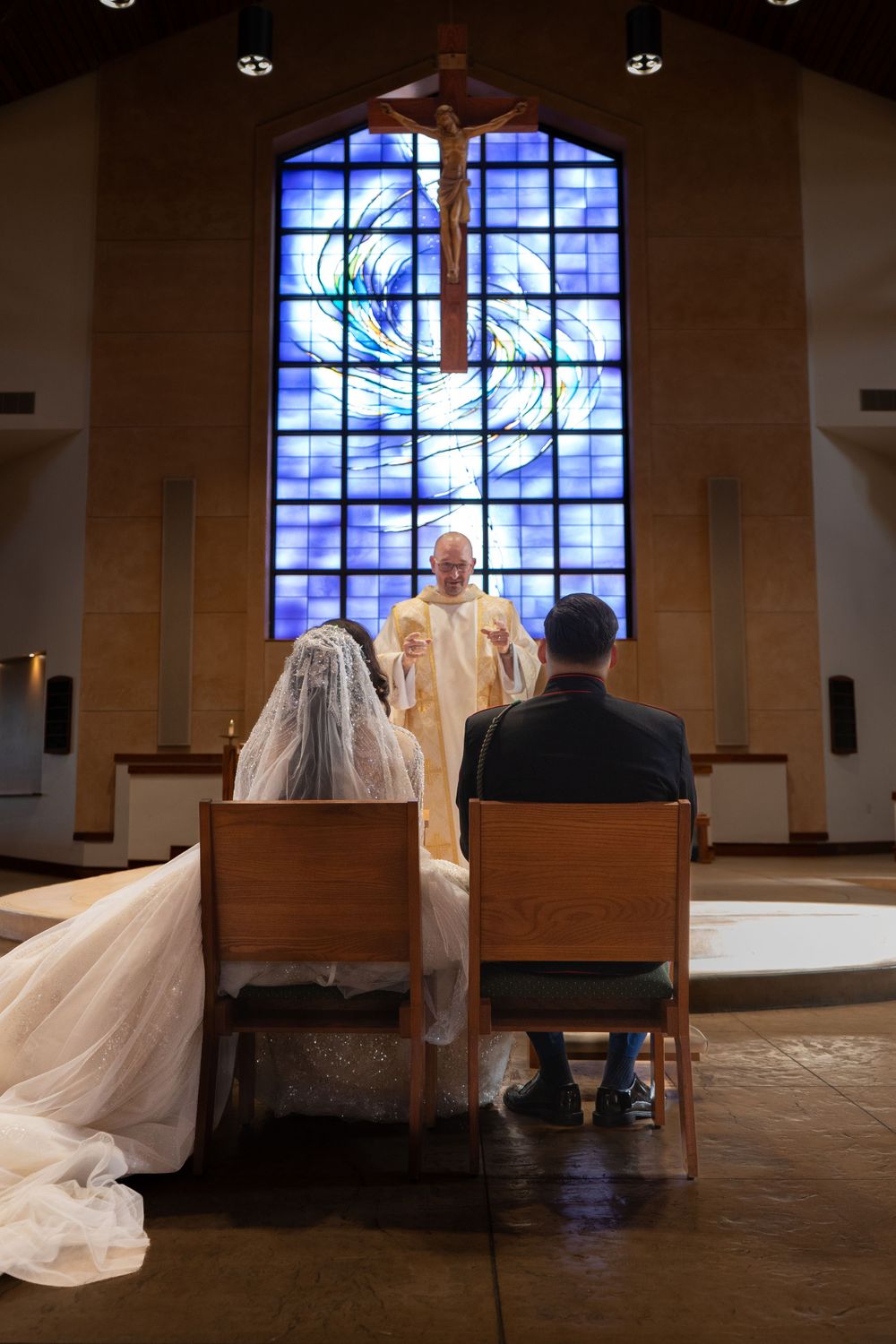 Bride and groom seated in church pews with dramatic stained glass windows in background.
