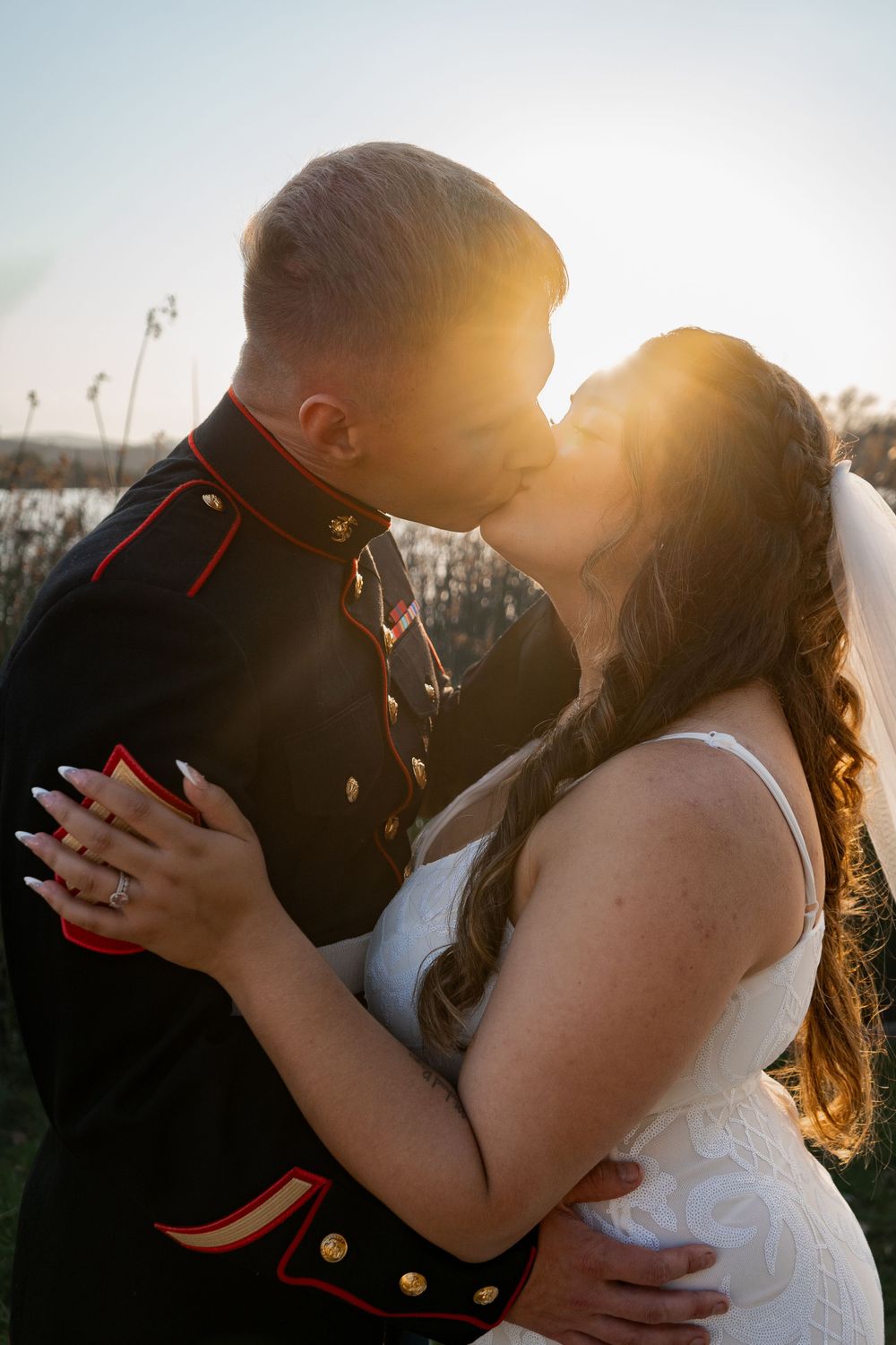A couple in marine dress uniform and white dress sharing a romantic sunset kiss.