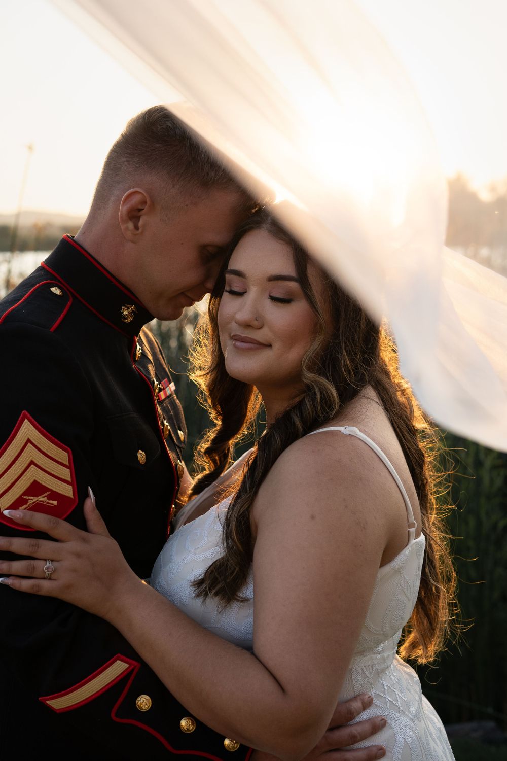 Newlywed couple embracing under a veil with golden sunlight streaming through.