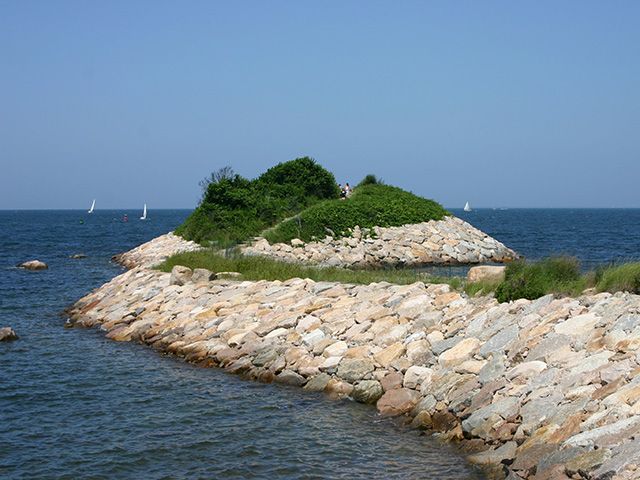 Rocky coastal outcrop with green vegetation and white sailboats visible in the blue ocean waters.