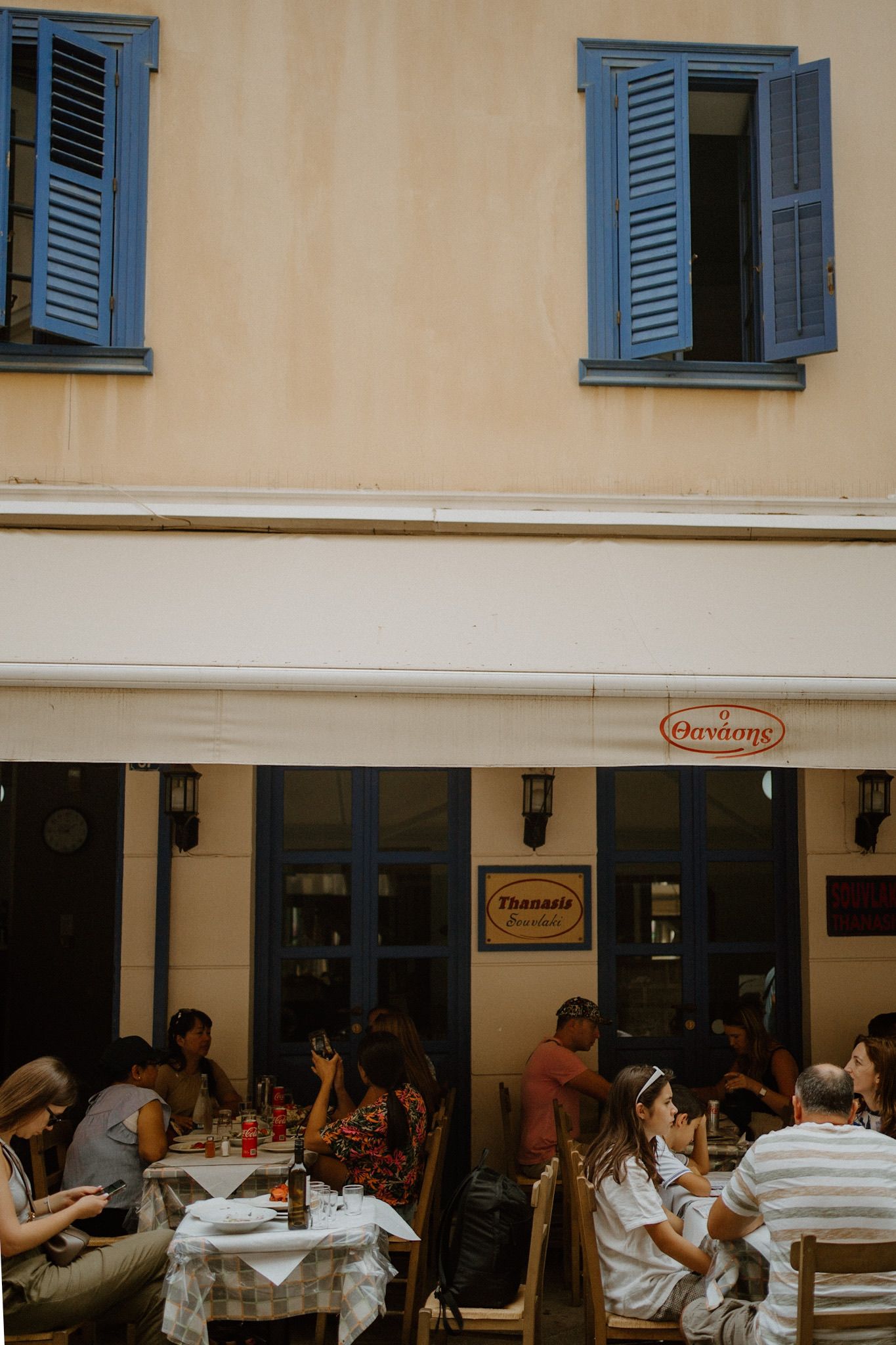 Diners enjoy outdoor seating at a rustic Italian restaurant with blue shuttered windows and cream colored walls.