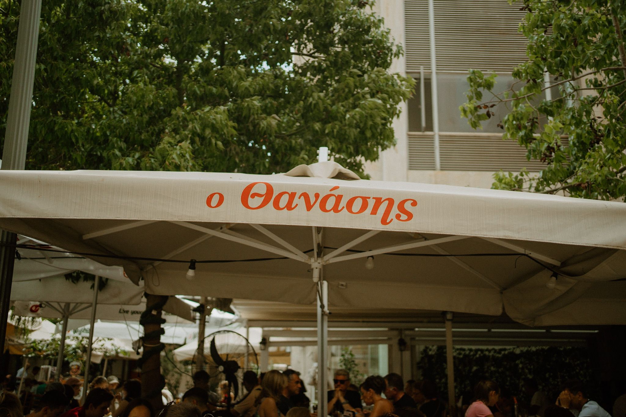 White restaurant umbrella with 'O Thanasis' text at an outdoor dining area surrounded by trees and patrons.