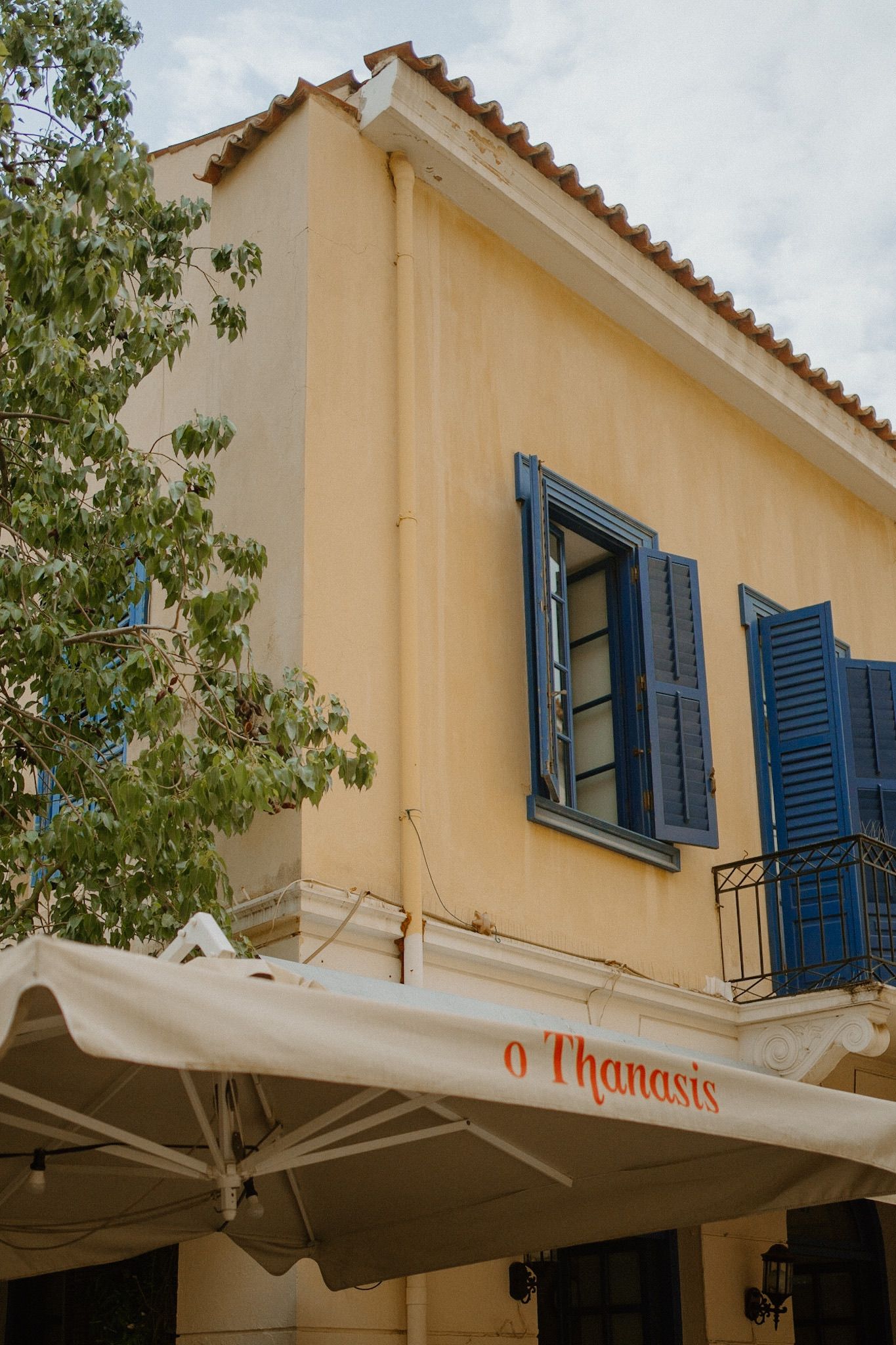 Mediterranean style building with cream colored walls, blue shuttered windows and a retractable awning for O'Thanasis cafe.