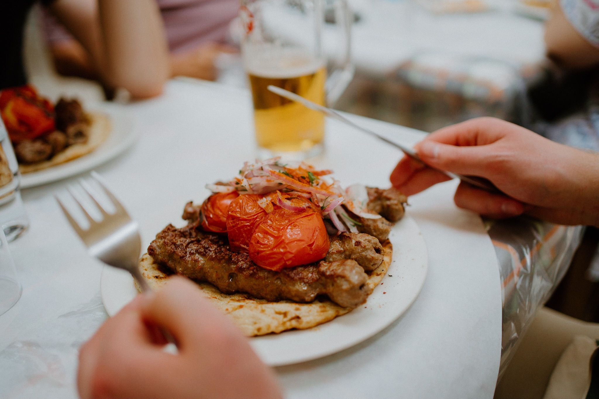 A juicy hamburger topped with caramelized onions and tomato being eaten at a restaurant table with a beer in the background.