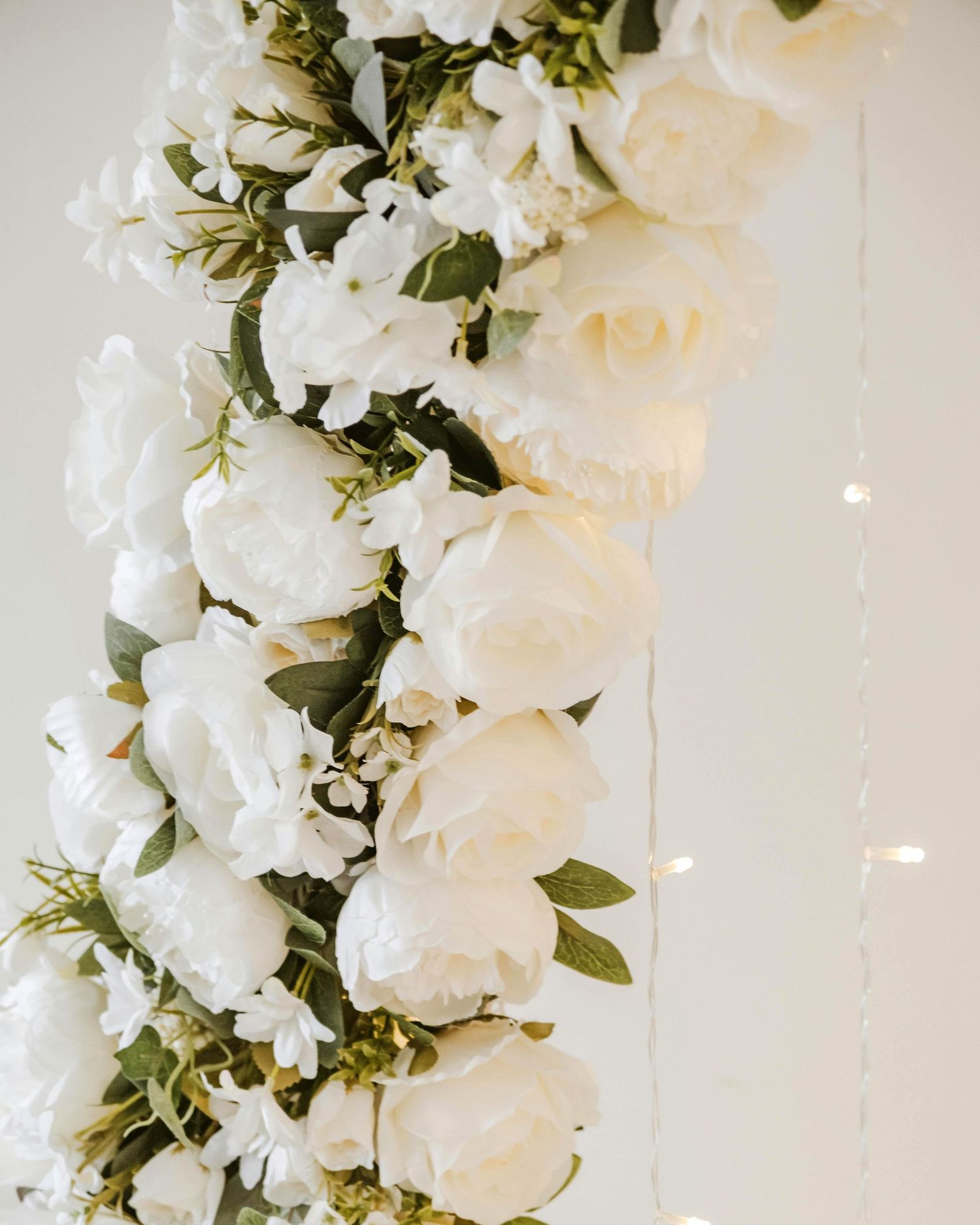 A vertical arrangement of white roses and greenery adorns a light-colored wall with subtle sparkles.