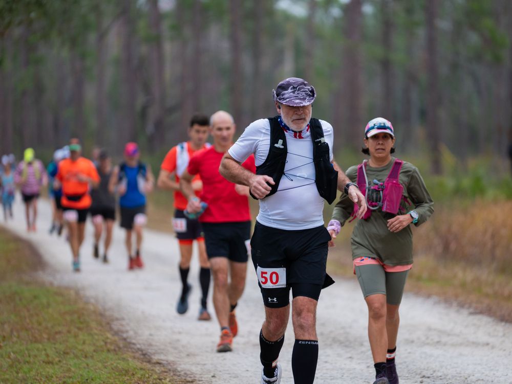 Athletes running along a white gravel path during a trail race through pine forest.