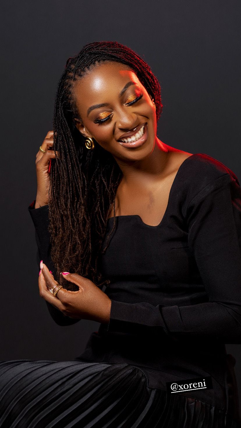 A joyful portrait with vibrant red eye makeup and long braids wearing a black outfit against dark background.