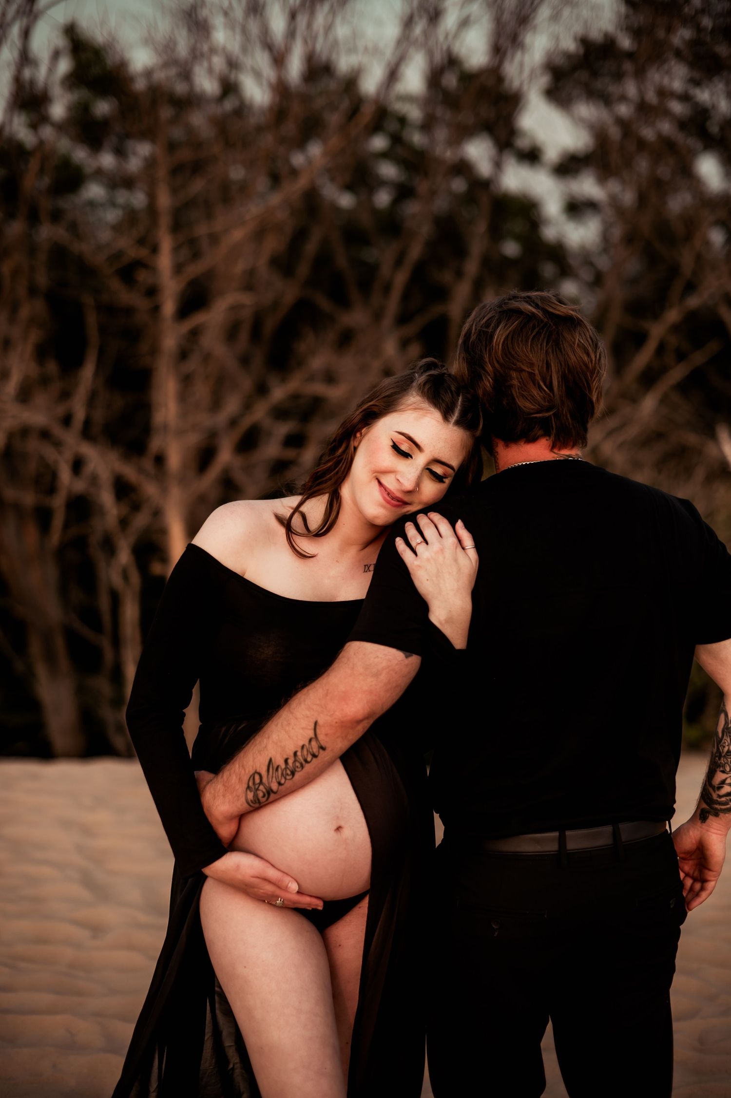A pregnant woman in a black dress embraces husband against a dark natural backdrop at sunset.