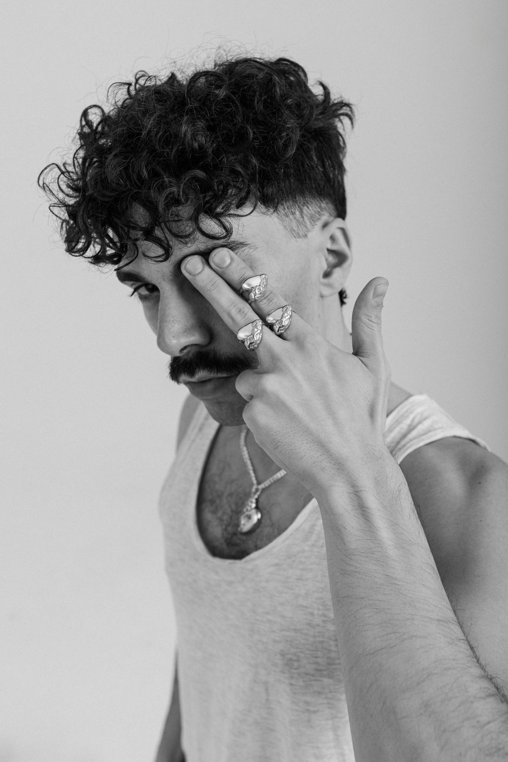 Black and white portrait of someone in a white tank top wearing rings and striking a dramatic pose with curly hair.