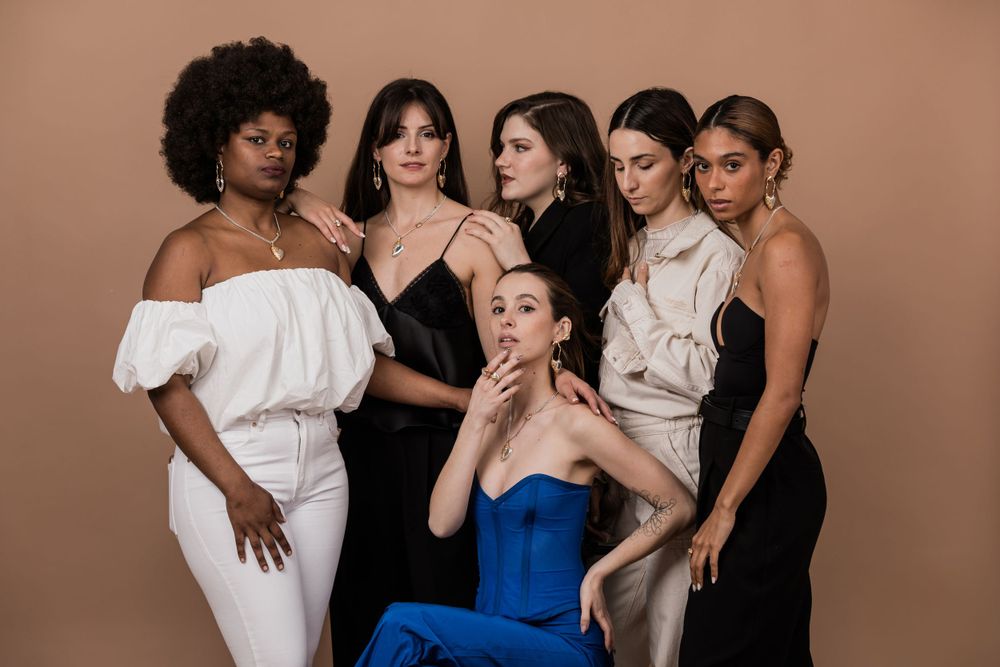 A group of six models in elegant evening wear pose together against a neutral brown backdrop.