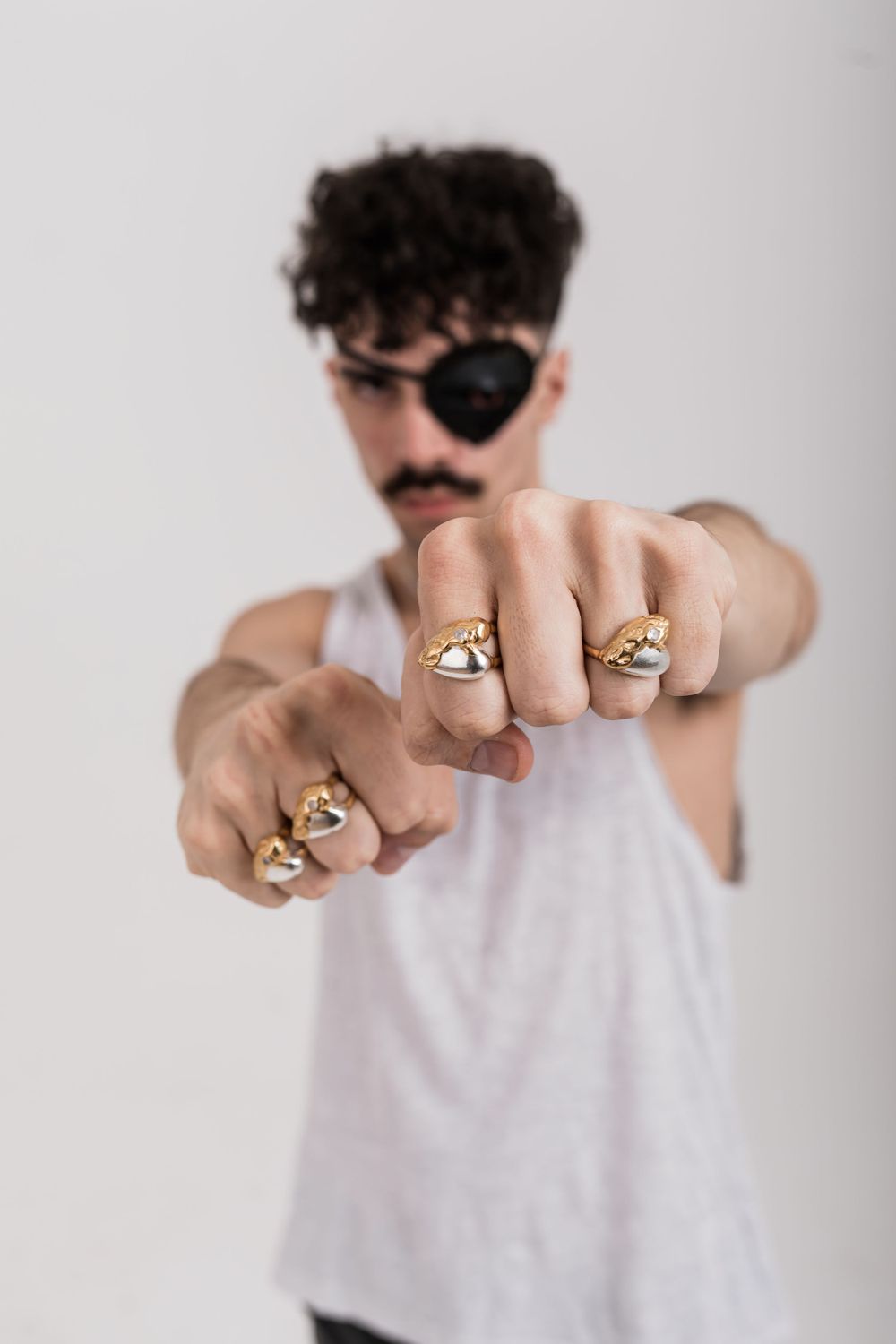 Person wearing gold rings on their fingers poses against white background with an eye patch and tank top.