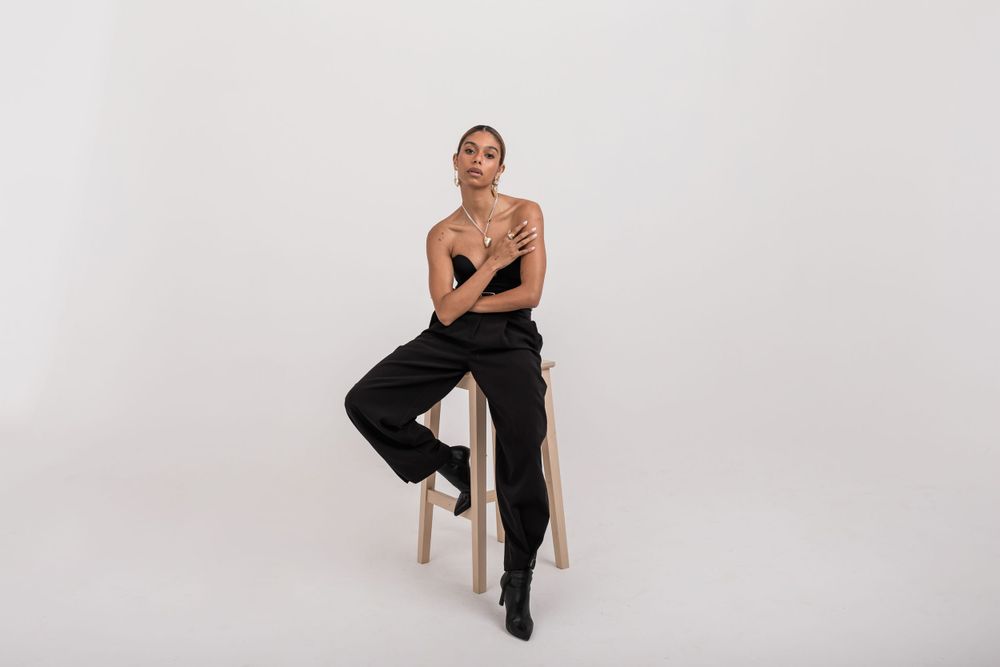 Minimalist portrait of someone seated on wooden stool wearing black pants against neutral gray backdrop.