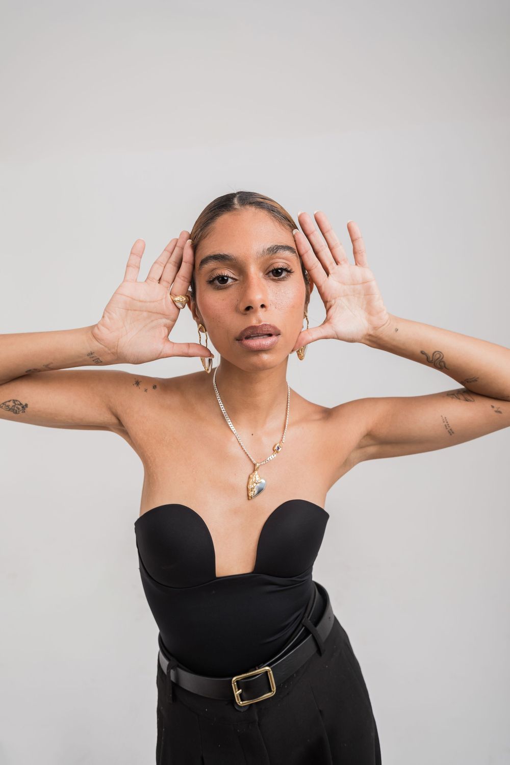 Fashion portrait showcasing a black strapless top with belt, crystal necklace and hands framing face against gray background.