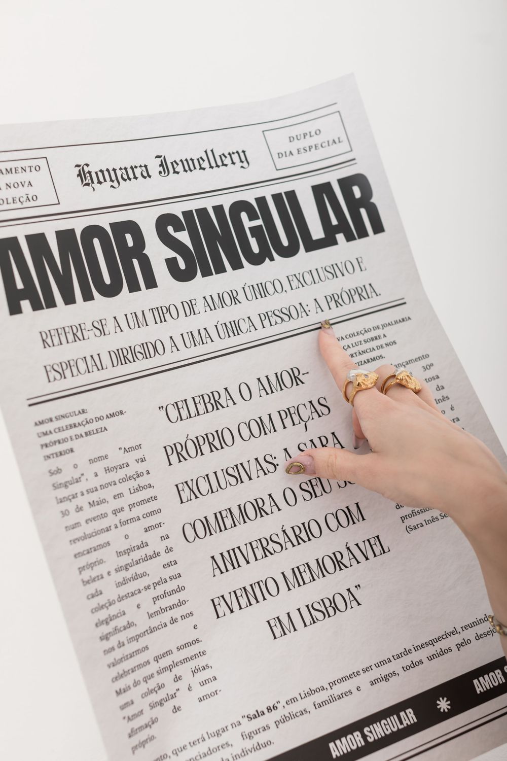 Hand wearing gold ring points at text on newspaper titled 'AMOR SINGULAR' with jewelry branding at the top.