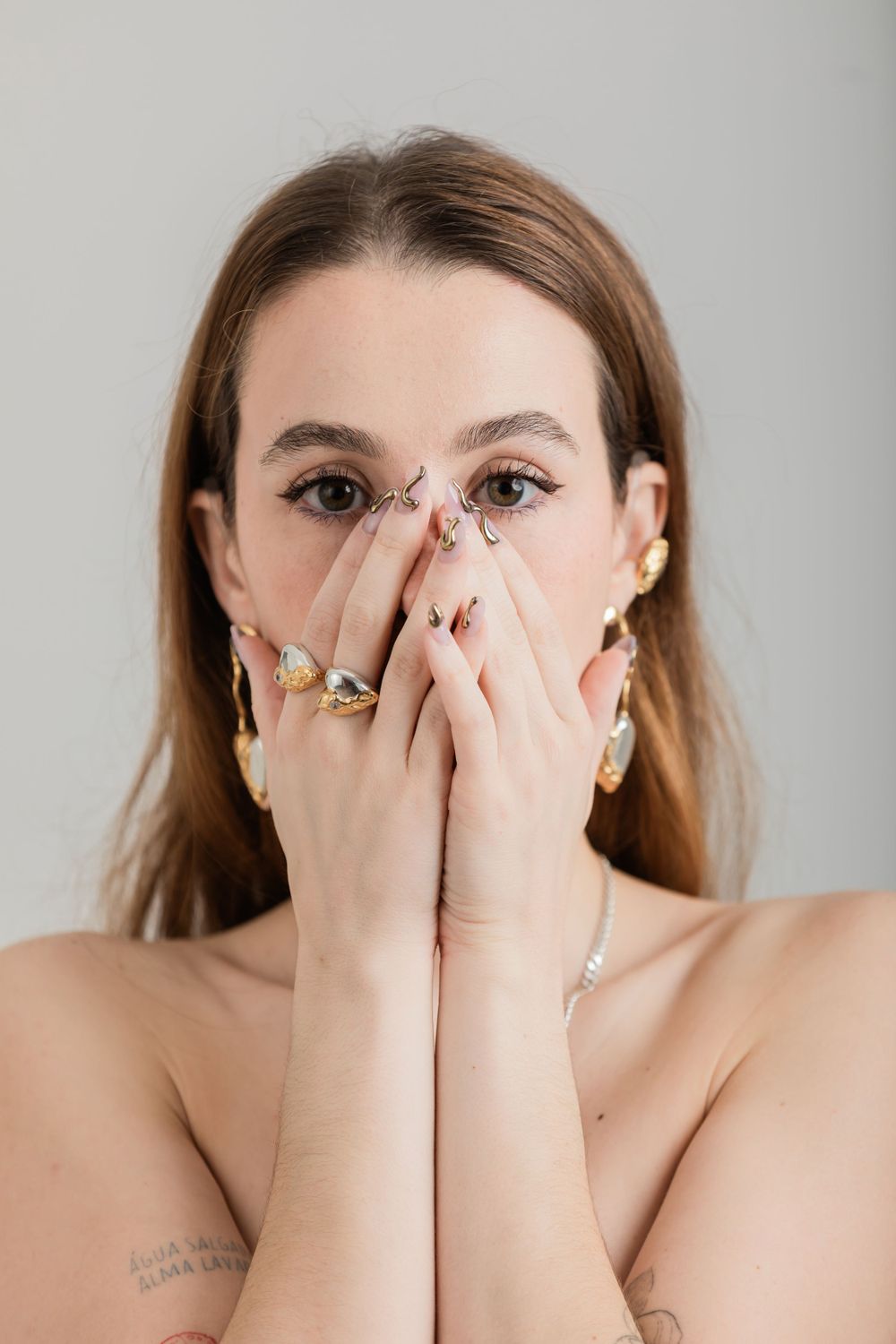 Person with brown hair wearing multiple rings covers their mouth with both hands against a light background.