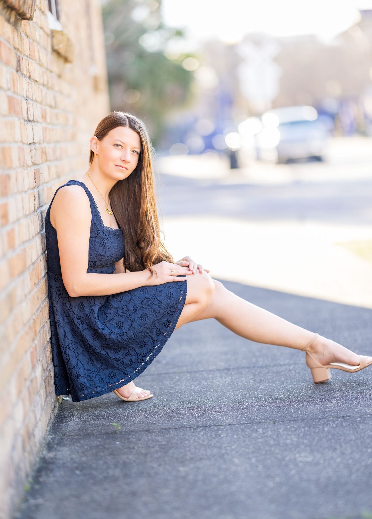 A person in a navy blue lace dress sits against a brick wall on a sunny day with cars visible in the background.