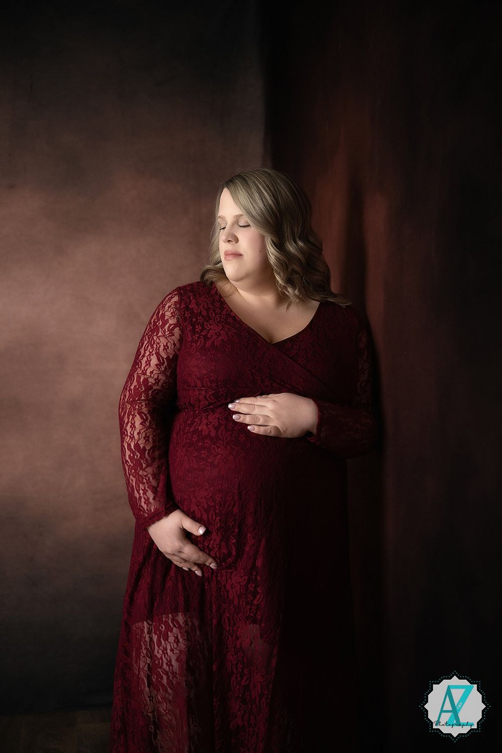 A maternity portrait featuring a burgundy lace dress against a moody dark background with dramatic lighting.