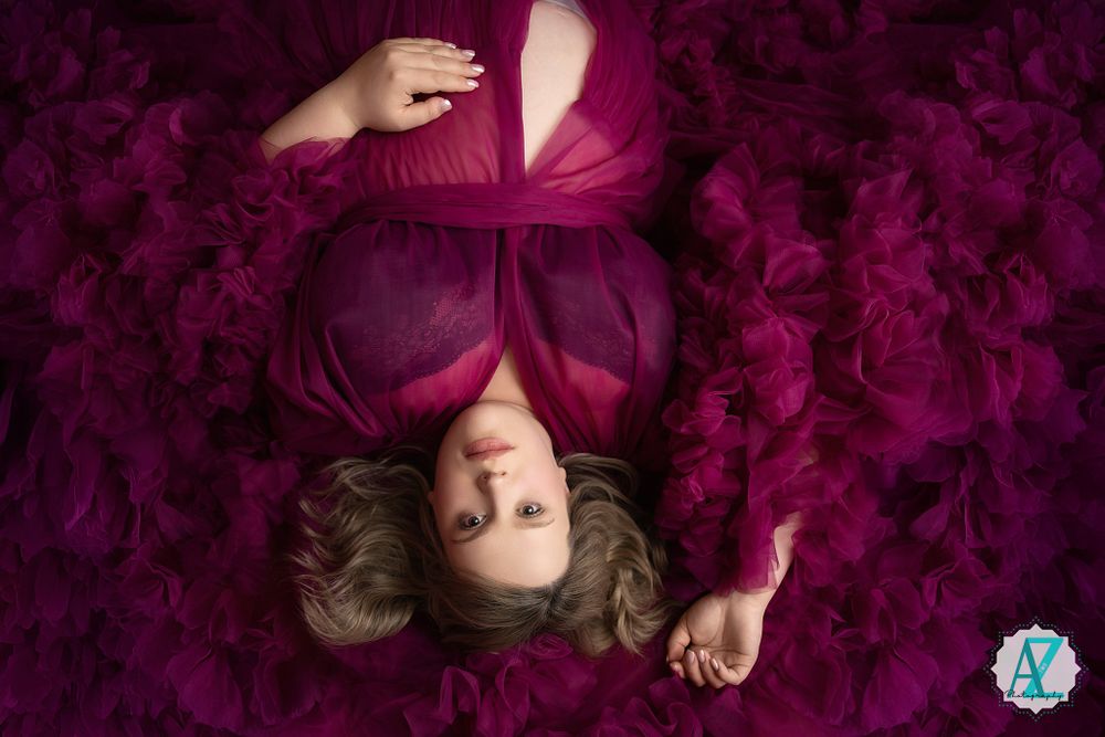 Overhead view of subject lying on purple textured fabric wearing a flowing burgundy dress posed with outstretched arms.