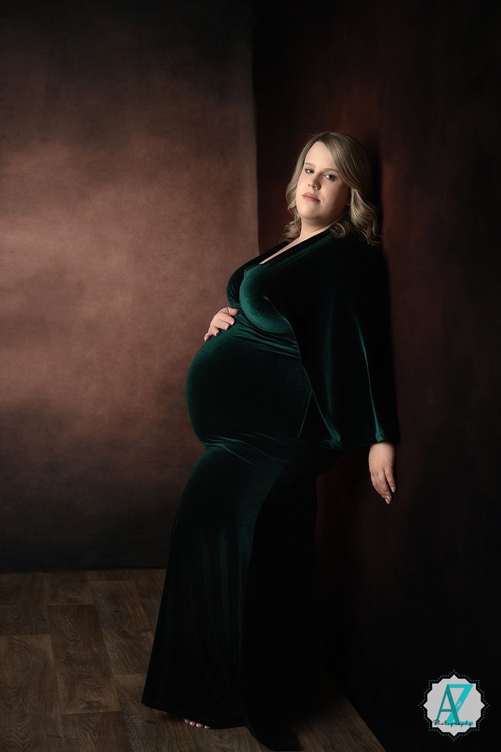 Elegant maternity portrait featuring a dark emerald green velvet gown against a moody brown studio backdrop.