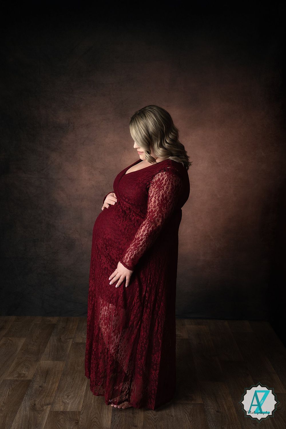 Elegant maternity portrait showing a woman in a burgundy lace gown posing against a dark moody backdrop.