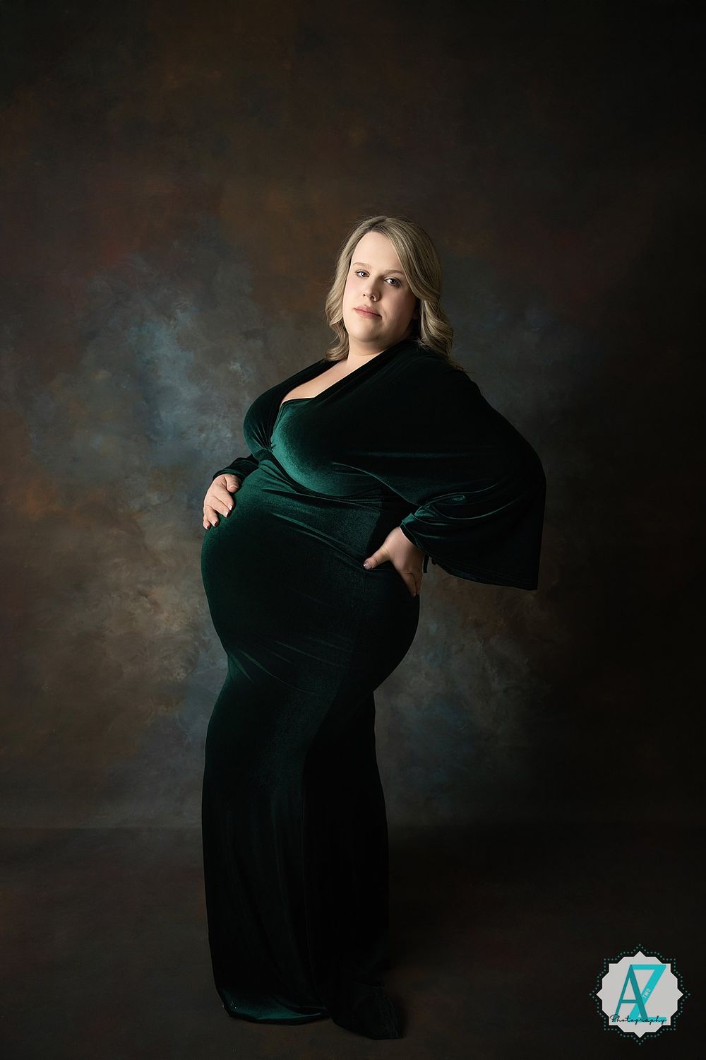 Maternity portrait with emerald green velvet gown against dark moody background with dramatic studio lighting.