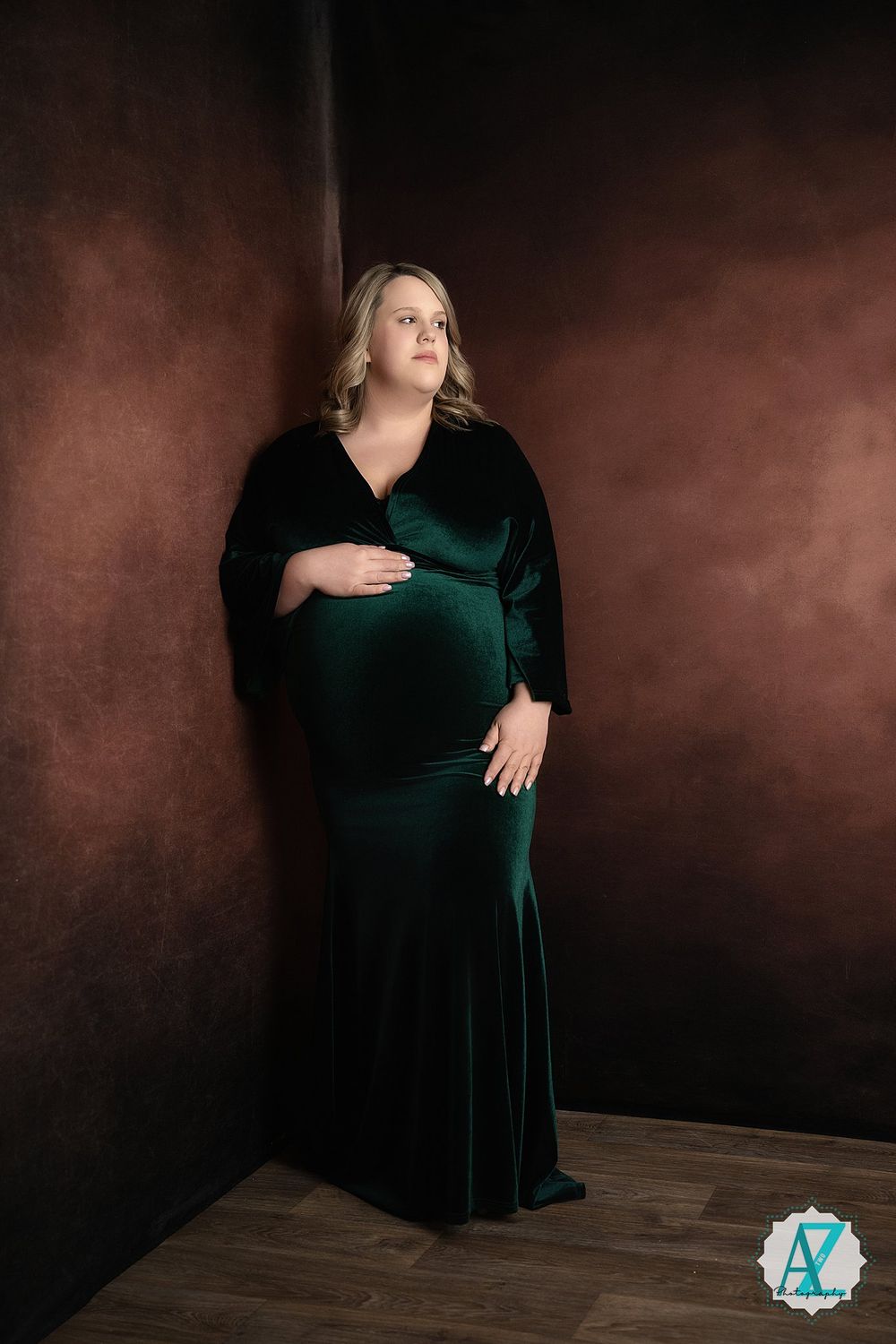 Elegant maternity portrait in emerald green velvet gown against moody brown backdrop with hardwood flooring.